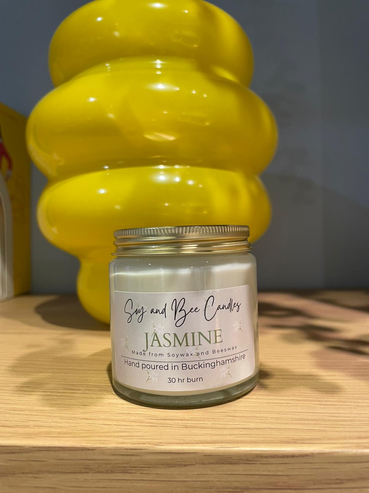 Jar of 'Jasmine' candle from Say and Bee Candles on a wooden surface with a yellow decorative object in the background.