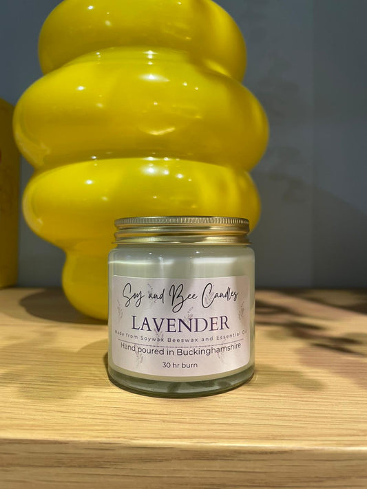 Lavender candle in a glass jar with a gold lid on a wooden surface, with a yellow decorative object in the background.